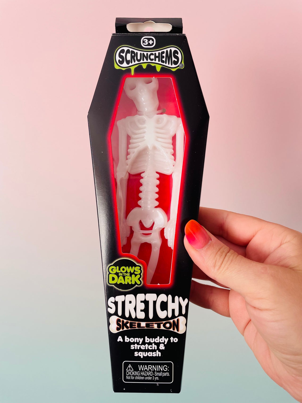 A hand holds a packaged Scrunchems Stretchy Skeleton toy in a coffin-shaped box against a pink-to-blue gradient background. The box is black with a clear window showing a white skeleton figure inside, and text on the package reads 'Scrunchems,' 'Stretchy Skeleton,' and 'Glow in the Dark.