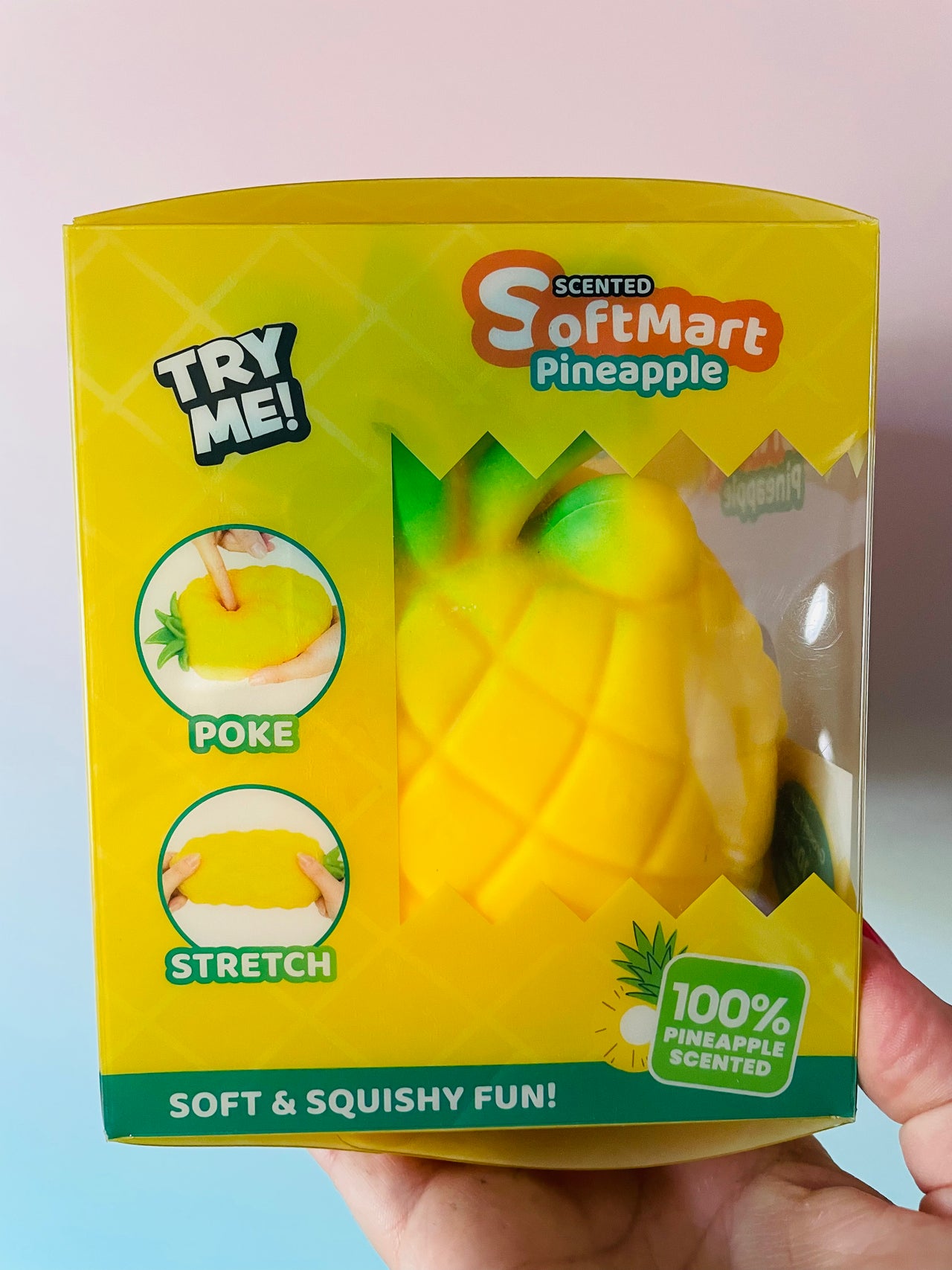 A hand holding a yellow SoftMart Pineapple-shaped squishy toy in its retail packaging, with a clear window showing the product inside. The box features product details and illustrations.