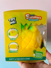 A hand holding a yellow SoftMart Pineapple-shaped squishy toy in its retail packaging, with a clear window showing the product inside. The box features product details and illustrations.