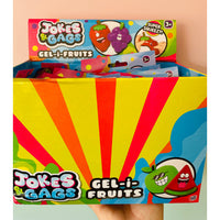 A hand holding a large box of Jokes Gags Gel-i-Fruits, showing the colorful packaging with red, yellow, and orange sunburst patterns and cartoon fruit illustrations. The top of the box displays individual packet packs.
