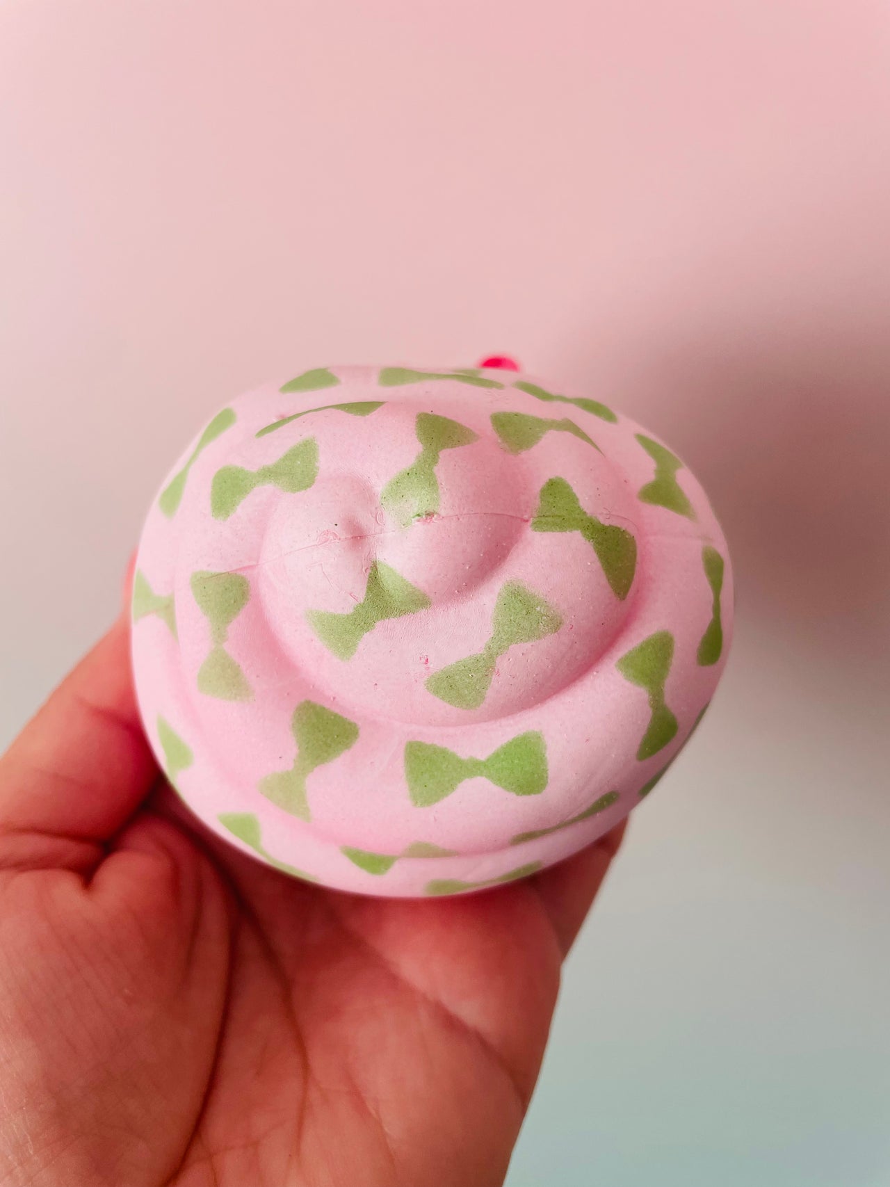 A hand holding a round, pink squishy toy with a pattern of green bow-tie shapes, set against a soft pink and light blue gradient background.