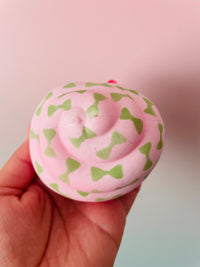 A hand holding a round, pink squishy toy with a pattern of green bow-tie shapes, set against a soft pink and light blue gradient background.