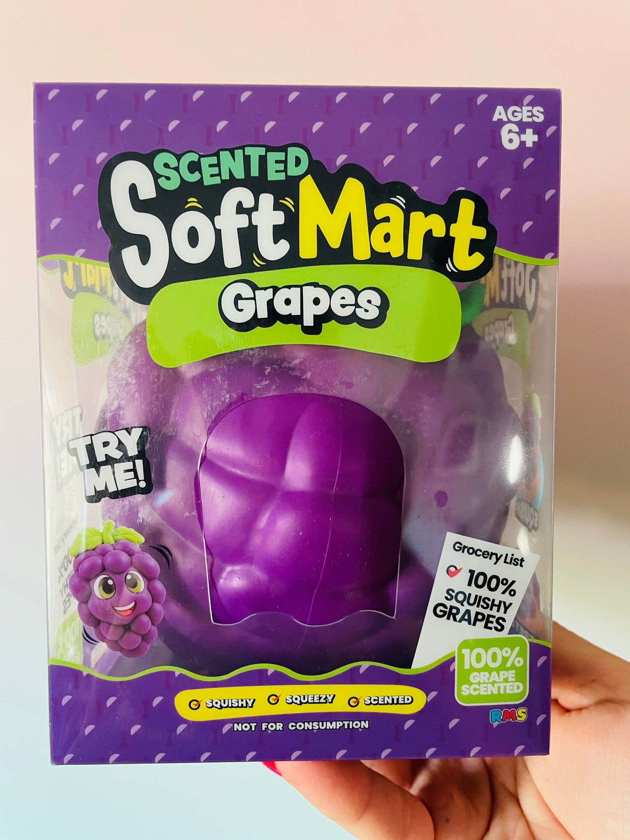 A hand holding a package of Scented Soft Mart Grapes toy. The purple packaging features a clear window showing the squishy purple grape-shaped toy inside.