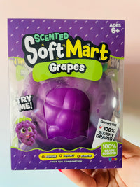 A hand holding a package of Scented Soft Mart Grapes toy. The purple packaging features a clear window showing the squishy purple grape-shaped toy inside.