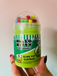 A hand with dark nail polish holds a clear plastic tub filled with bright green slime. The tub has a green and white striped label reading 'HELLO KITTY AND FRIENDS SLIME POT SURPRISE' with Hello Kitty graphics, and a clear dome lid filled with small colorful pom-poms.
