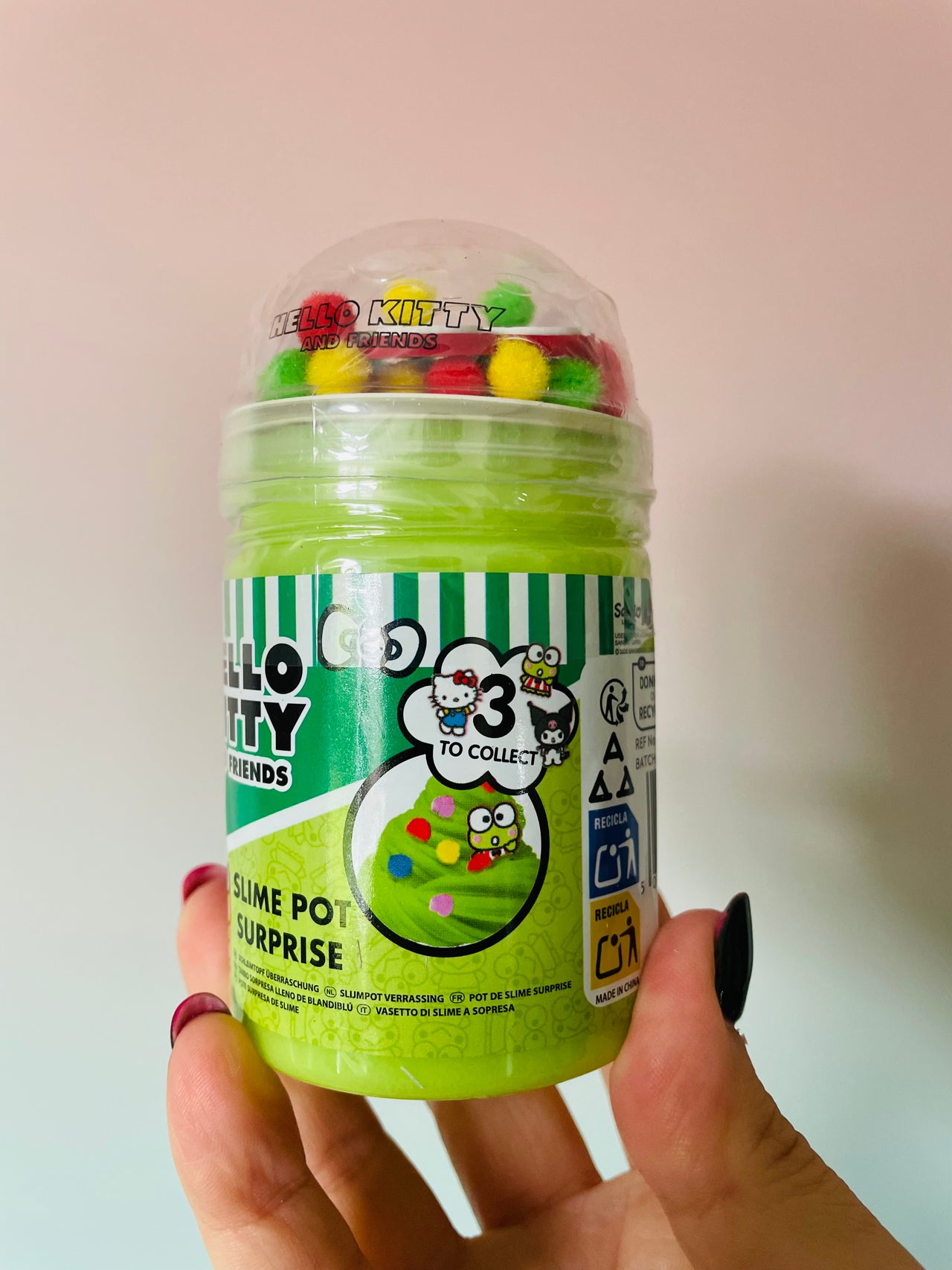 A hand holding a Hello Kitty Friends 'Slime Pot Surprise' collectible slime jar. The product is a bright lime green translucent slime inside a clear plastic jar with a clear dome lid filled with colorful round candies.